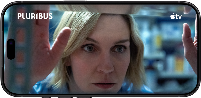iPhone 18 Pro vises i liggende format, på skjermen er det et nærbilde av en kvinne som ser ut gjennom et vindu. Serietittel: Pluribus
