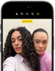 iPhone 18 Pro i fargen svart vises forfra, to modeller poserer for kameraet, for å vise Center Stage-frontkameraet